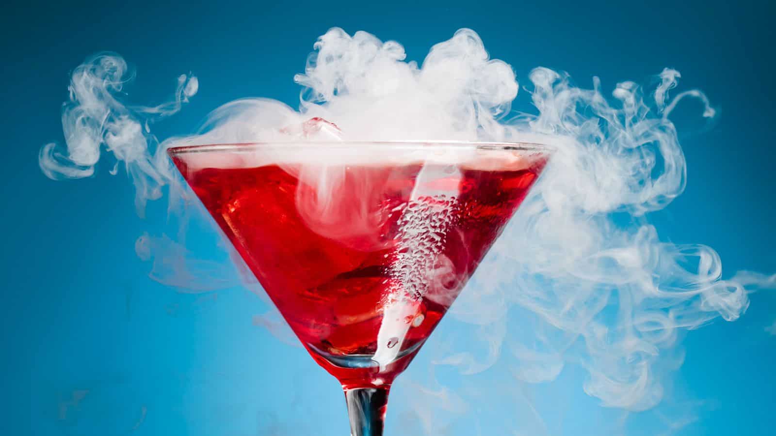 A cocktail glass filled with a red liquid emits swirling white smoke against a blue background. Bubbles are visible in the drink, enhancing the smoky effect.