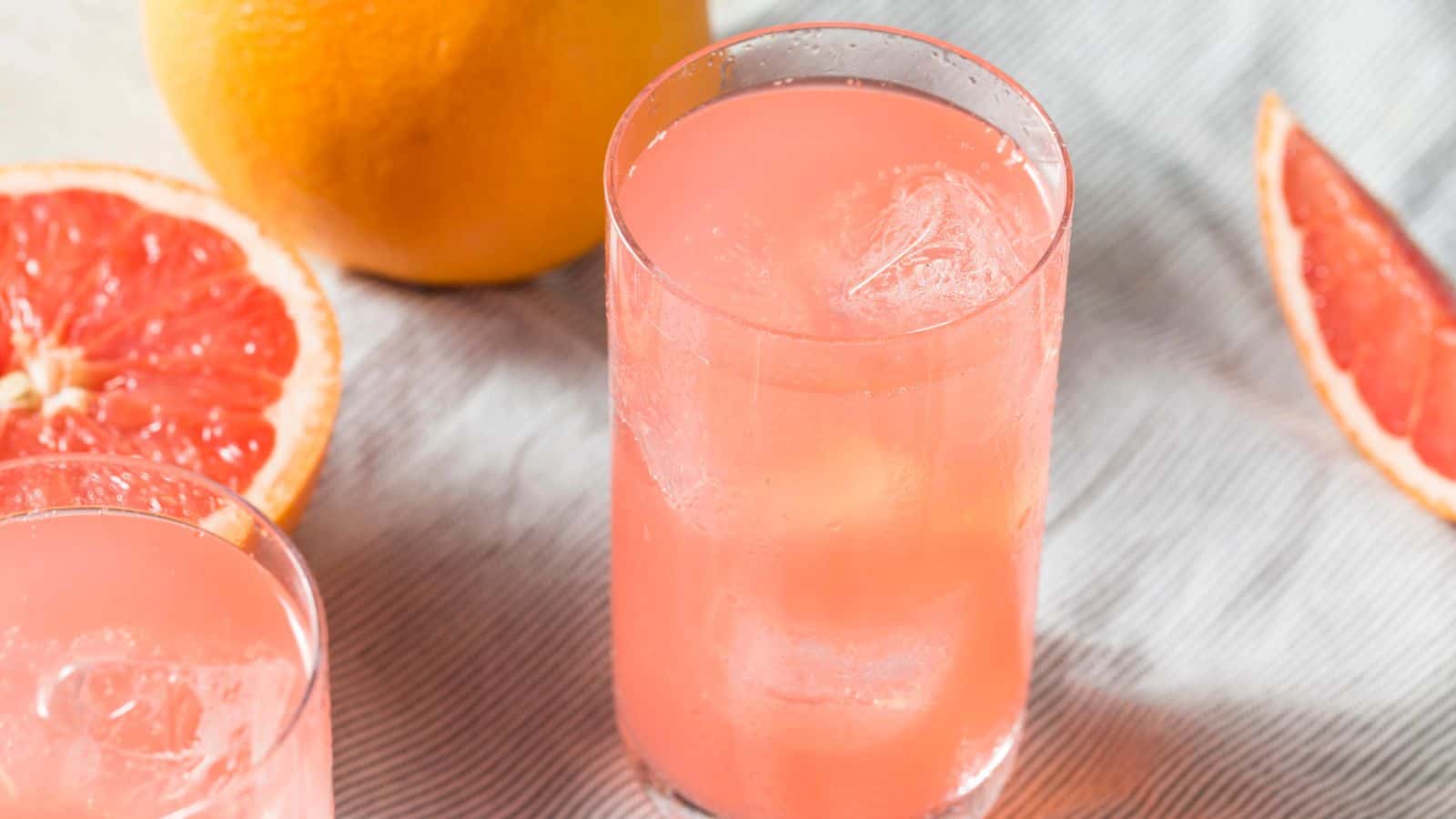 A clear glass filled with pink grapefruit juice, containing ice cubes, is placed on a striped cloth. A whole grapefruit and several grapefruit slices are adjacent to the glass. Another partially visible glass is on the left.
