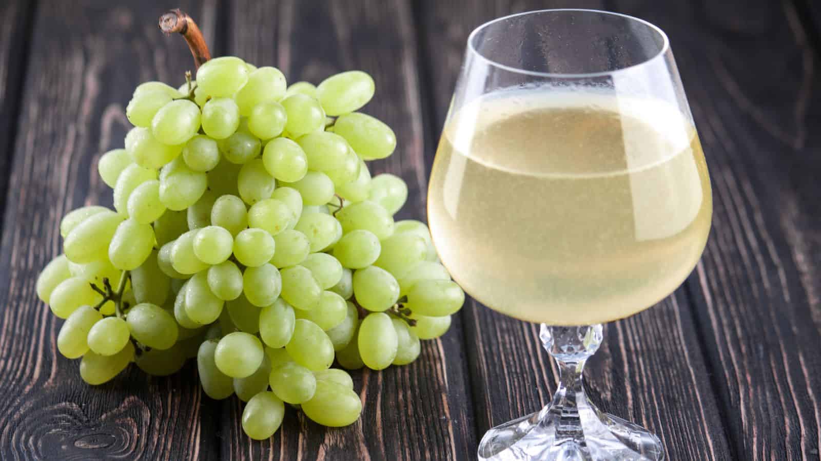 A bunch of green grapes lies next to a glass filled with a clear, light yellow liquid on a dark wooden surface.