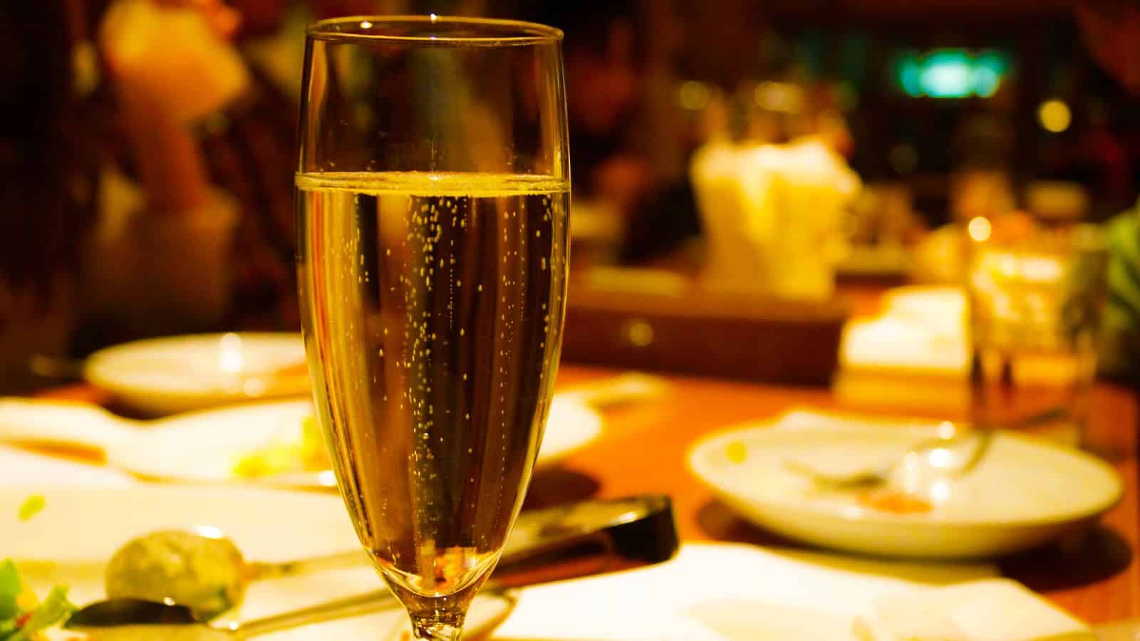 A tall glass of champagne sits on a dining table set with white plates, cutlery, and a blurred background of diners. The glass is filled with bubbling champagne, capturing a warm, inviting ambiance.