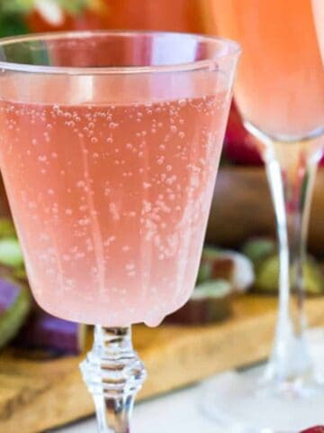 A glass filled with a bubbly pink drink is in focus. In the background, there are two more glasses with similar drinks. Cut lemons, strawberries, and flowers are also visible on a wooden board.