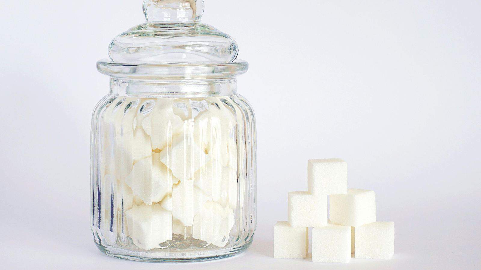 A glass jar filled with white sugar cubes is placed next to a small pyramid of sugar cubes. The jar has a lid and sits on a plain white surface against a light background.