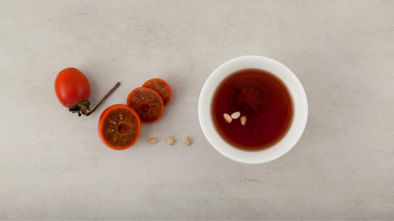 A whole persimmon and three sliced persimmon pieces are on the left, with some seeds placed in a row. On the right, there is a white cup filled with a brown liquid. The background is a light grey surface.