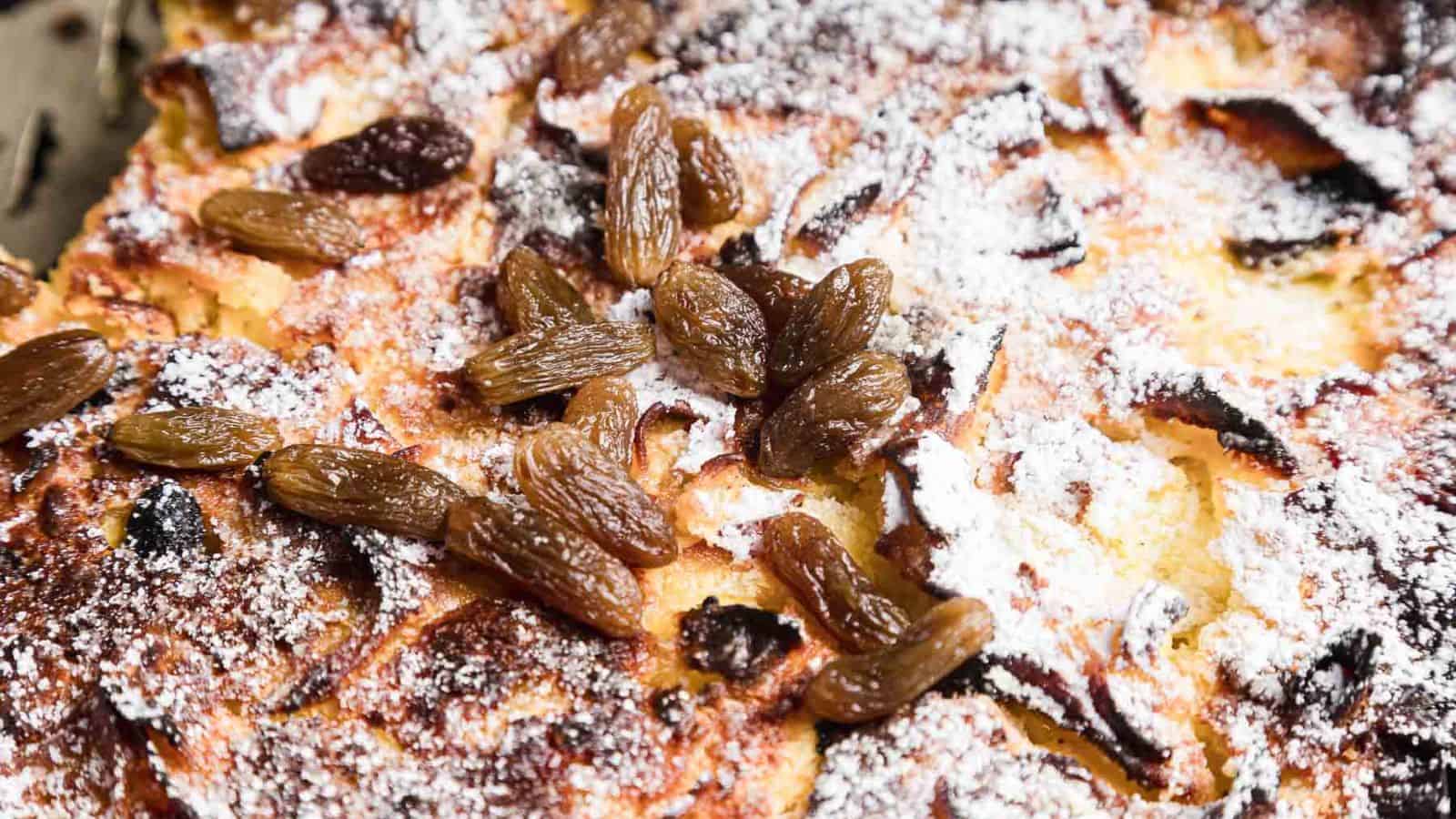 A slice of bread pudding on a decorative plate with a fork, topped with whipped cream. A baking dish with more bread pudding and a small bowl of cream with a spoon are in the background. A brown cloth is partially visible on the side.