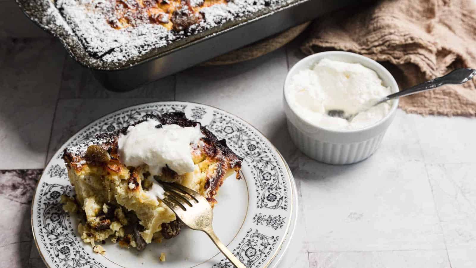A slice of bread pudding on a decorative plate with a fork, topped with whipped cream. A baking dish with more bread pudding and a small bowl of cream with a spoon are in the background. A brown cloth is partially visible on the side.