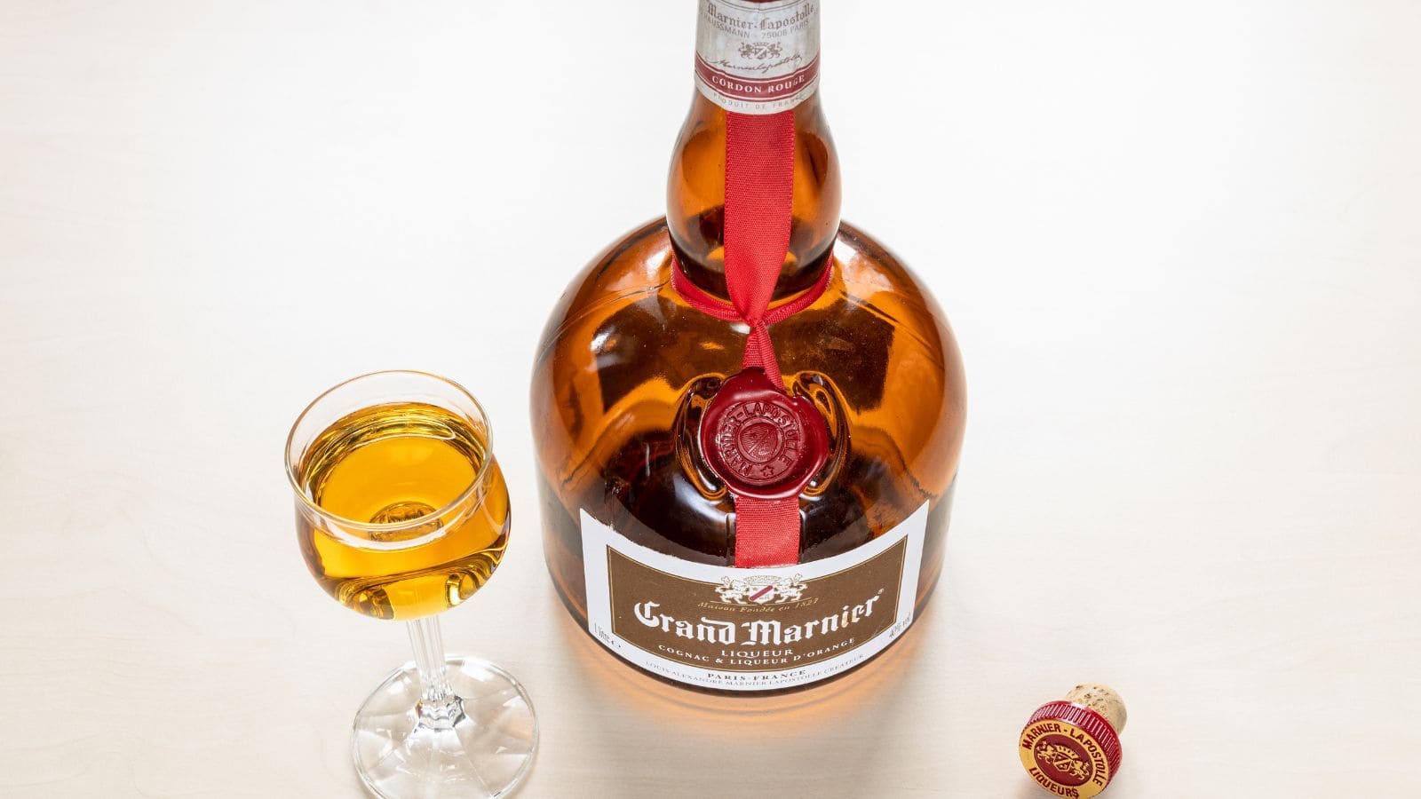 A large bottle of Grand Marnier liqueur with a red ribbon and wax seal sits on a light surface. Next to the bottle is a filled cordial glass and a detached cork with a red top on the table.