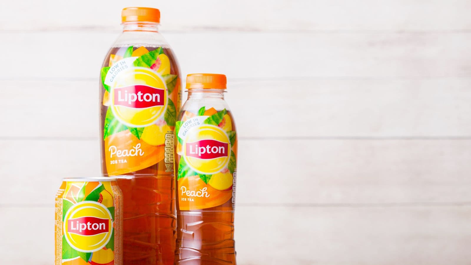 Three Lipton peach iced tea containers are displayed against a light wooden background. The containers include a can, a medium-sized bottle, and a large bottle, each with a yellow and orange design featuring peaches and tea leaves.