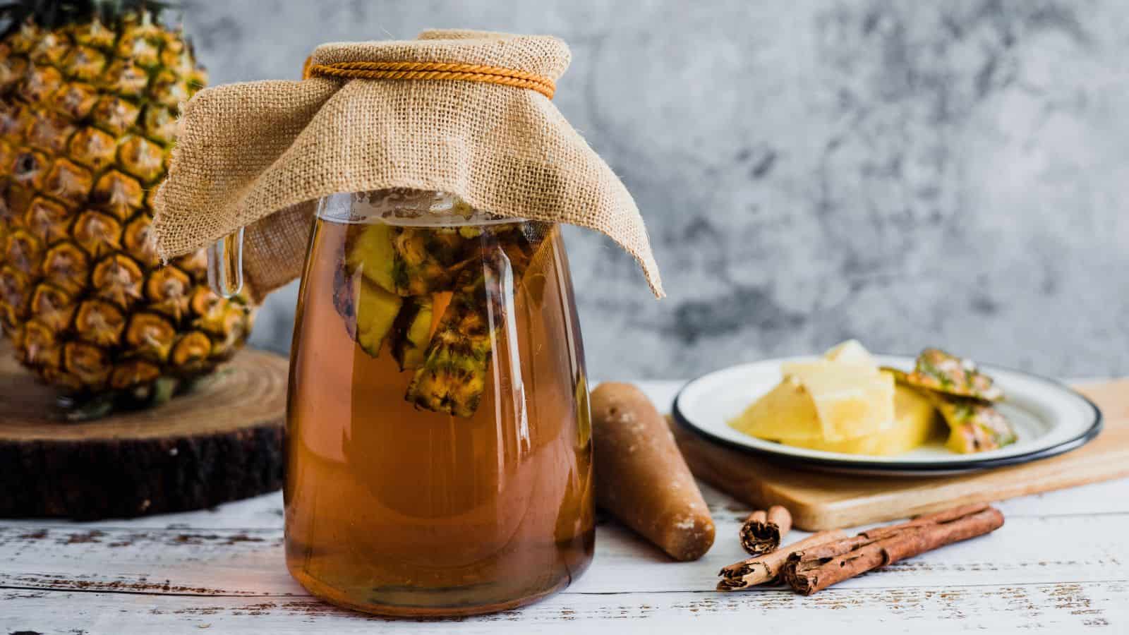 A glass jar with a burlap cover contains a brown liquid and pineapple chunks. It is placed on a wooden surface alongside a whole pineapple, a log slice, cinnamon sticks, a few spices, and a plate with cut pineapple pieces.