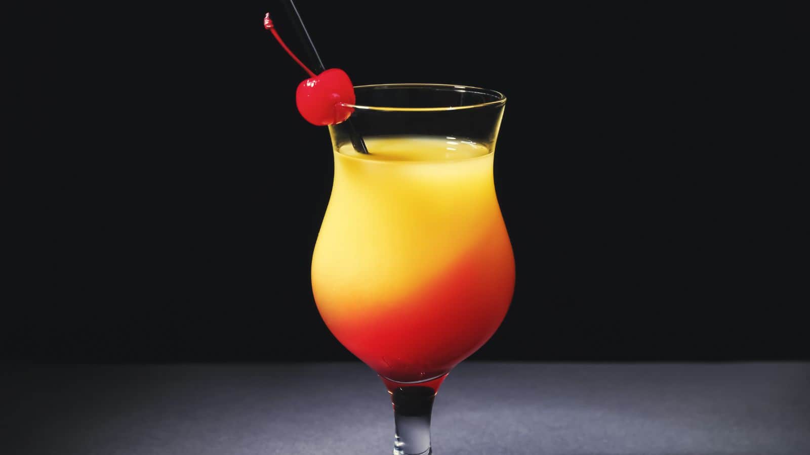 A vibrant cocktail in a curved glass shows a gradient from orange to red. A cherry and black straw garnish the drink. The background is dark, emphasizing the bright colors of the beverage.