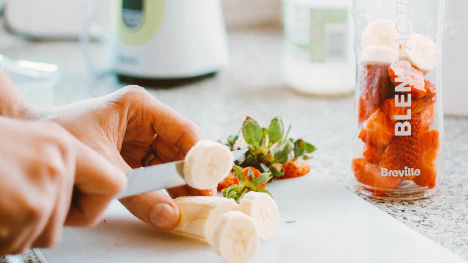 A person slices a banana with a knife on a white cutting board. Strawberries and banana slices are visible inside a blender cup labeled "Breville" on a countertop. A few green strawberry tops are on the cutting board.