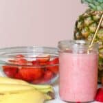 A glass jar filled with pink smoothie and a straw is surrounded by a pineapple, bananas, oranges, raspberries, and a bowl of sliced strawberries on a white surface against a light background.