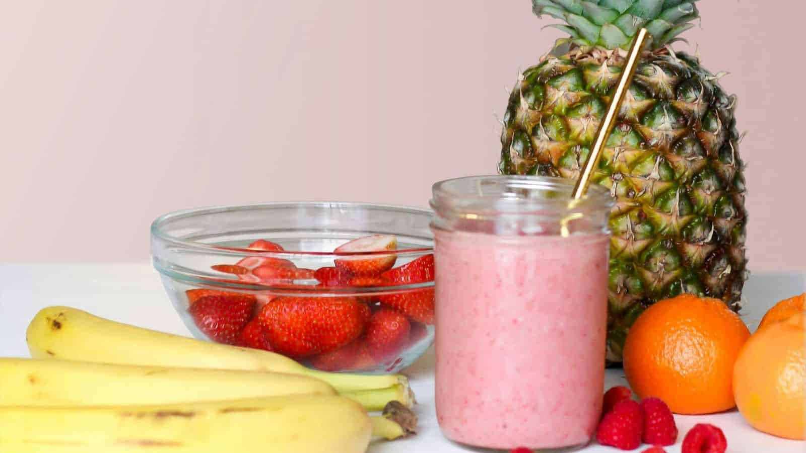A glass jar filled with pink smoothie and a straw is surrounded by a pineapple, bananas, oranges, raspberries, and a bowl of sliced strawberries on a white surface against a light background.