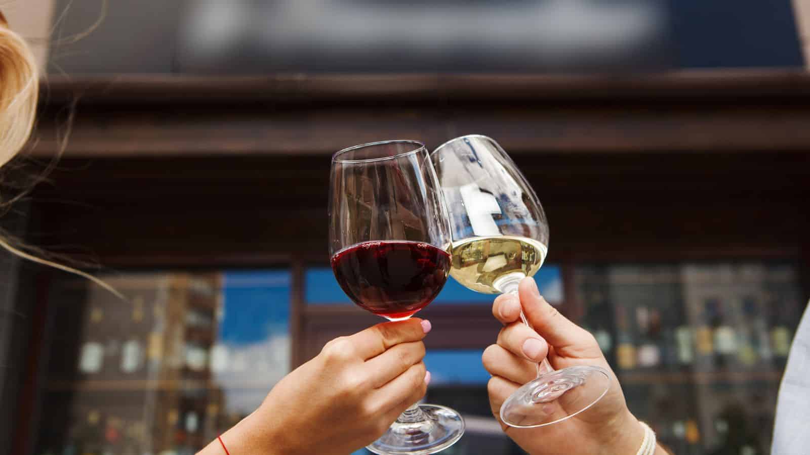 Two people clink glasses filled with red and white wine in an outdoor setting. The background shows a blurred building with glass windows displaying various wine bottles. One hand has a red bracelet.