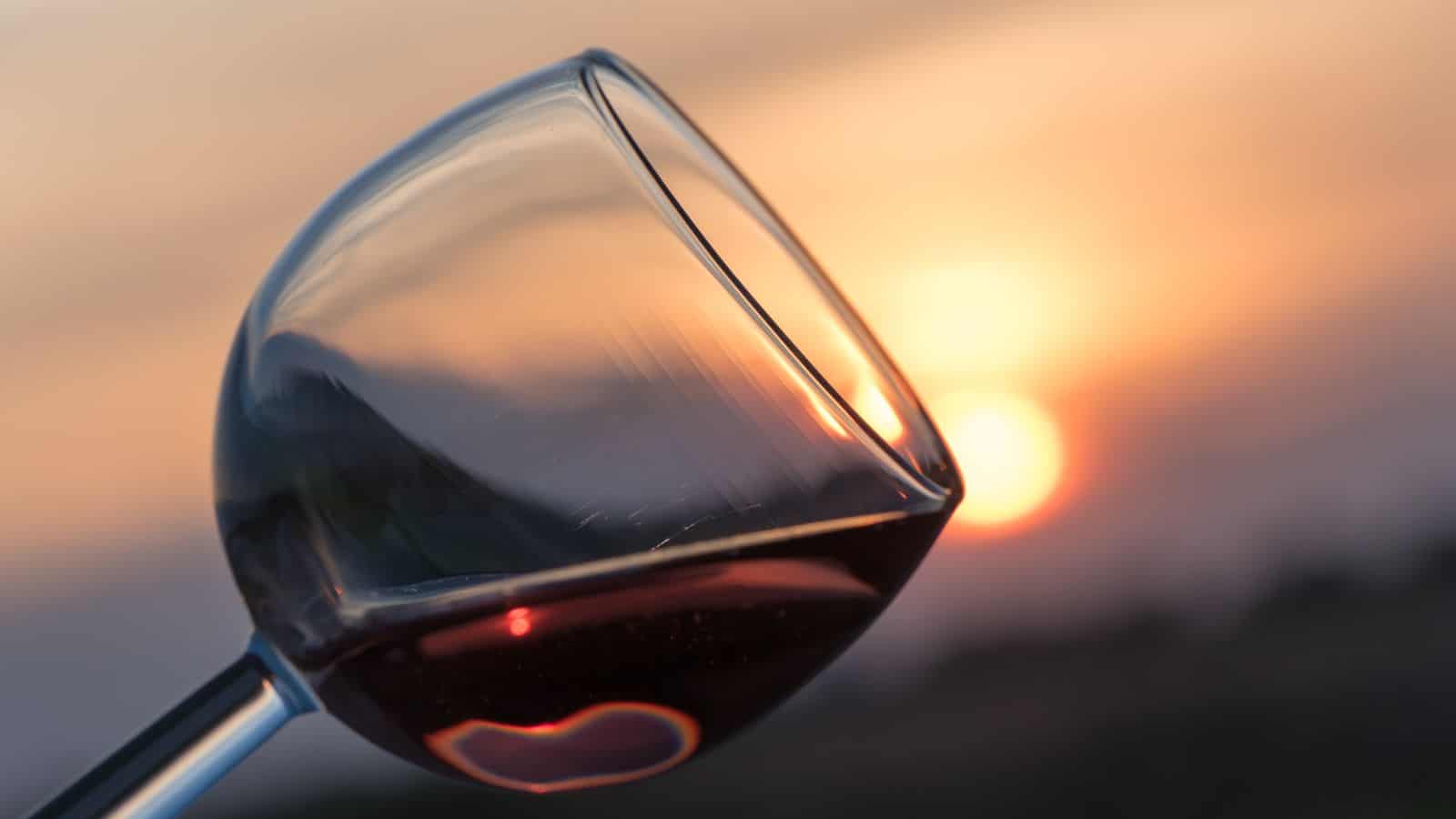 Close-up of a wine glass tilted against a sunset backdrop. The glass contains a small amount of red wine, and the warm hues of the sunset create a gradient of orange and pink in the background.