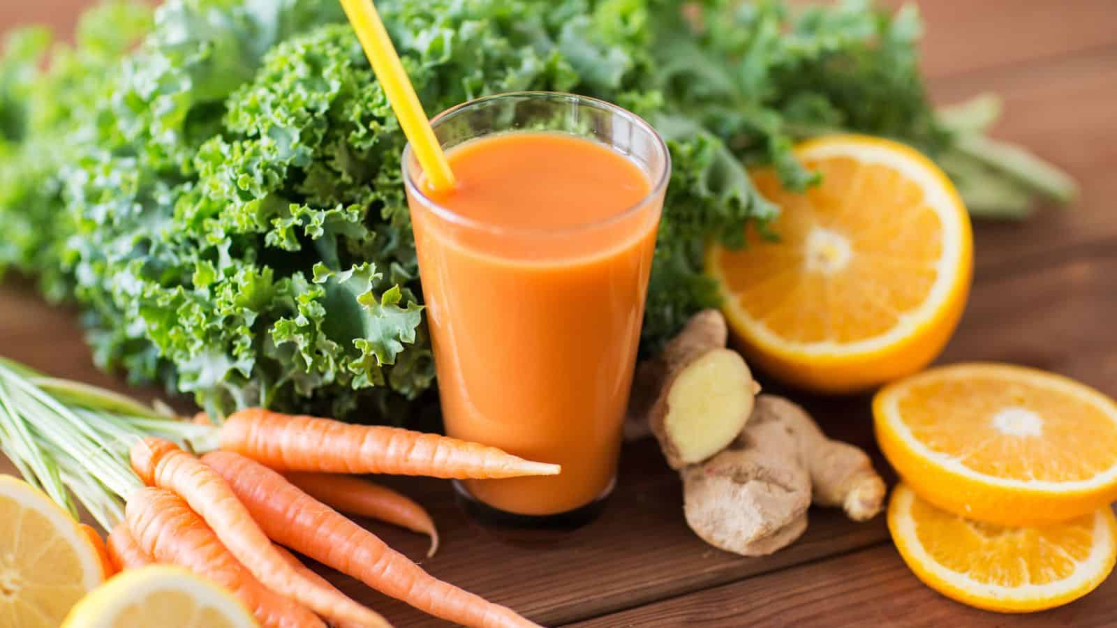 A glass of orange juice with a straw is surrounded by kale, sliced oranges, ginger root, and whole carrots on a wooden surface. The arrangement suggests fresh ingredients for a healthy drink.