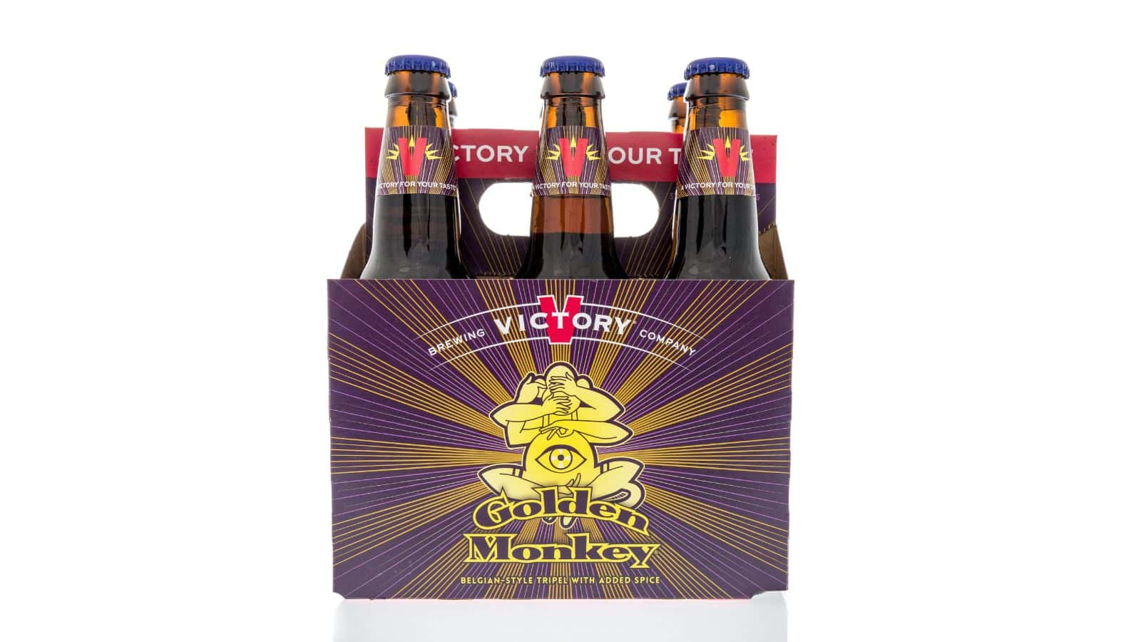 A six-pack of Golden Monkey beer bottles in a purple cardboard carrier with a yellow monkey design and text reading "Victory." The carrier has handles, and the bottle caps are blue. The background is white.