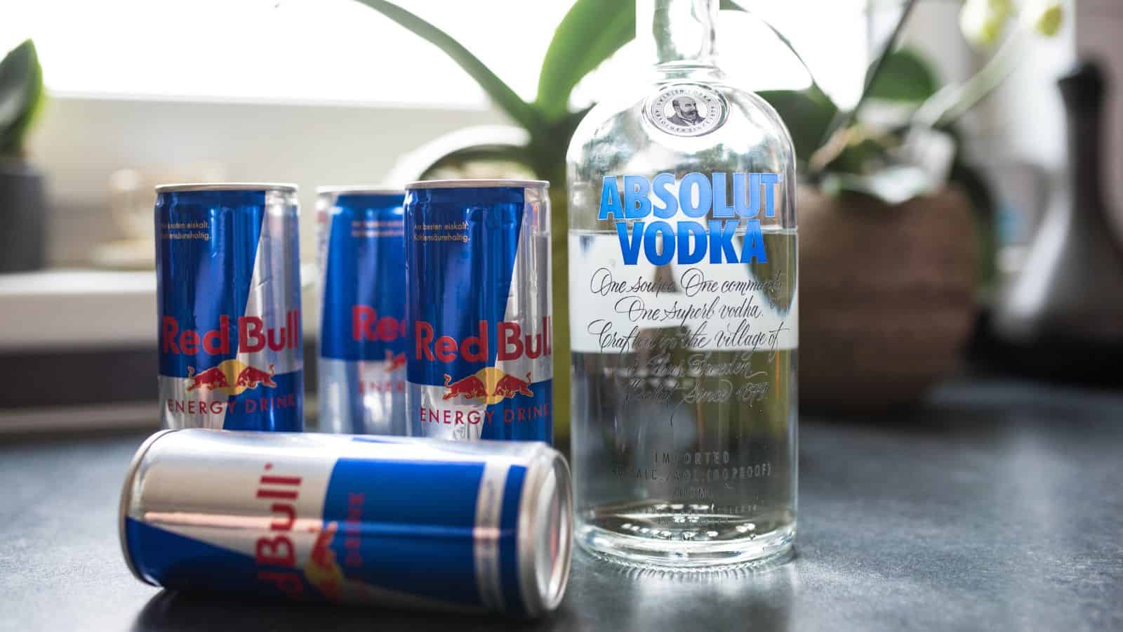 Four Red Bull energy drink cans are next to an Absolut Vodka bottle on a kitchen countertop with a plant in the background.
