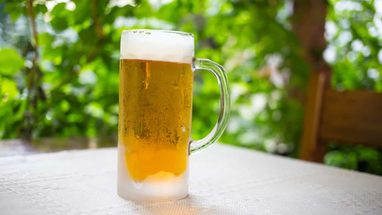 A frosty mug of beer with a foamy head sits on a white tablecloth. The background is lush with greenery, creating a natural outdoor setting.