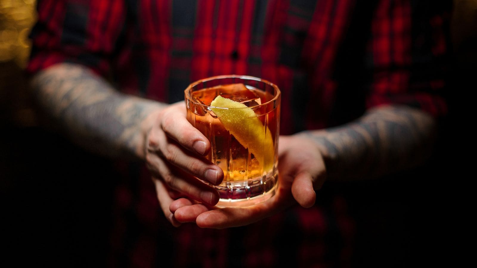 A person with tattooed arms holds a glass of amber-colored liquid with a slice of lemon peel in it. The person is wearing a red plaid shirt and the background is dark.