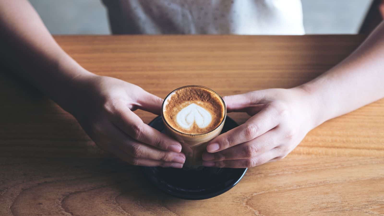 A person holds a small cup of coffee with heart-shaped latte art on top. The cup is on a black saucer placed on a wooden table. The person's hands are gently grasping the cup.