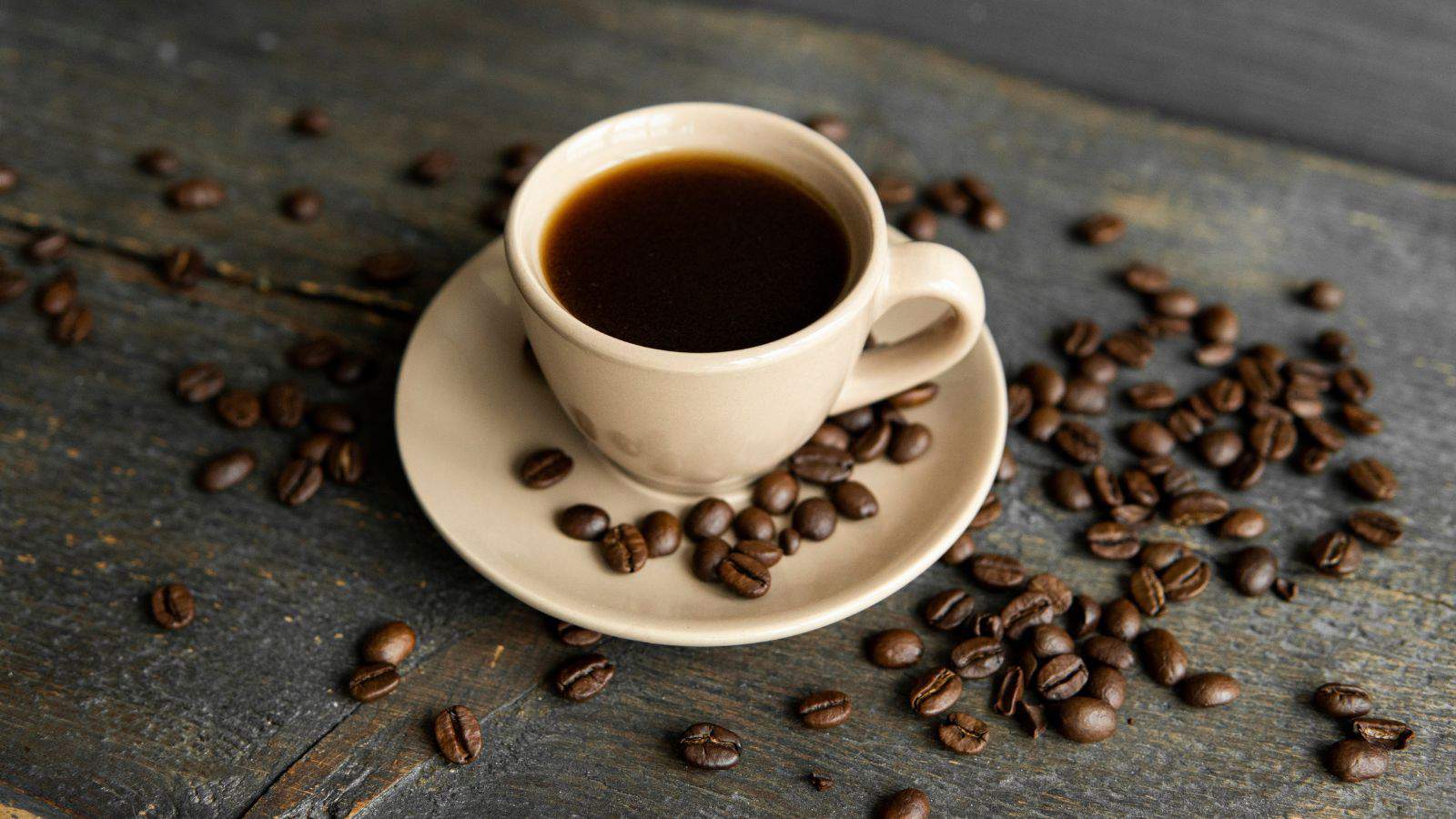 A white ceramic cup filled with dark coffee is placed on a matching saucer. Coffee beans are scattered around on a rustic wooden surface. The background is a dark, textured surface.