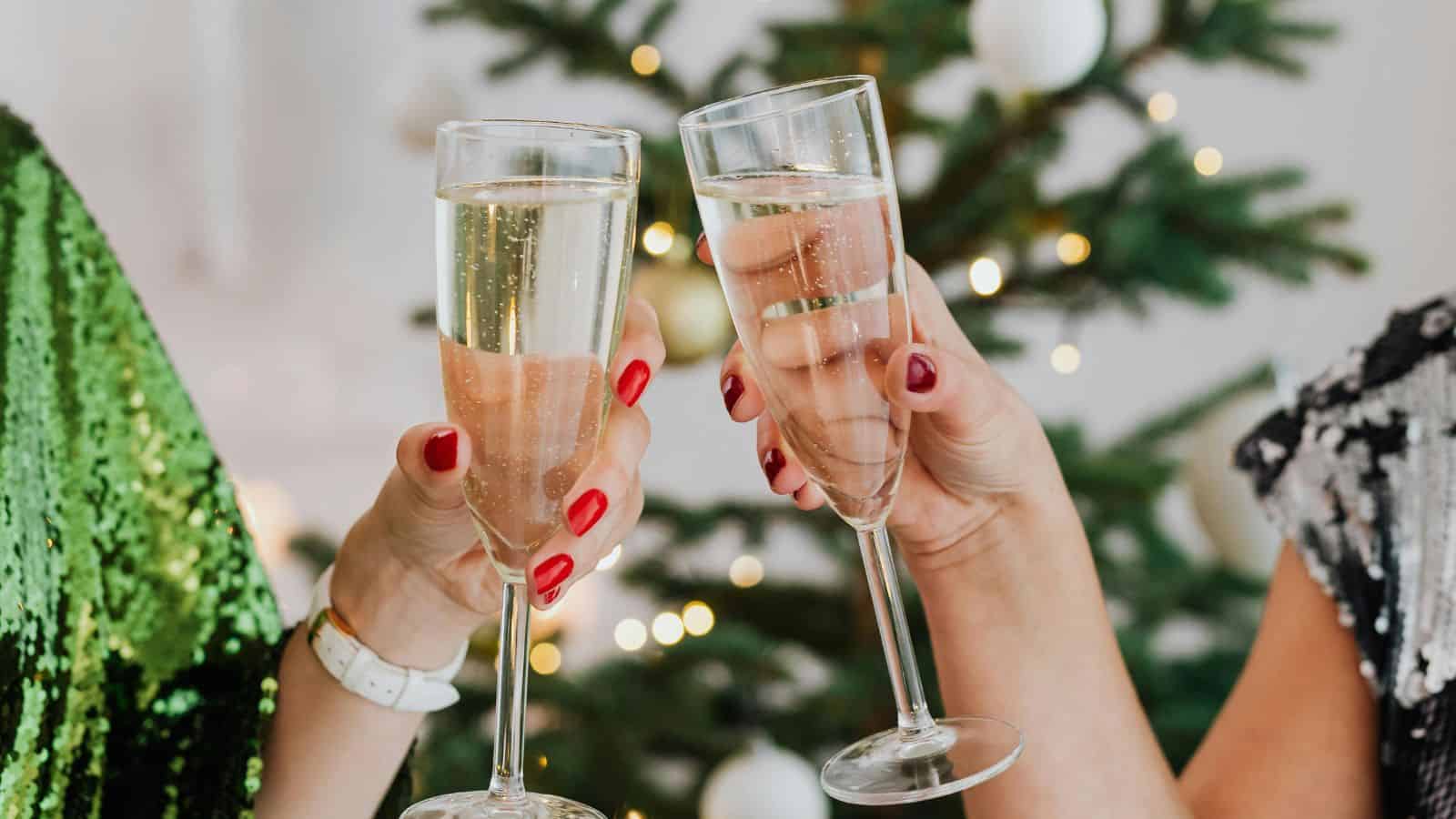 Two people clink champagne glasses in front of a decorated Christmas tree. The person on the left wears a green sequined garment, while the one on the right wears a silver sequined top. Each has a manicure in shades of red.
