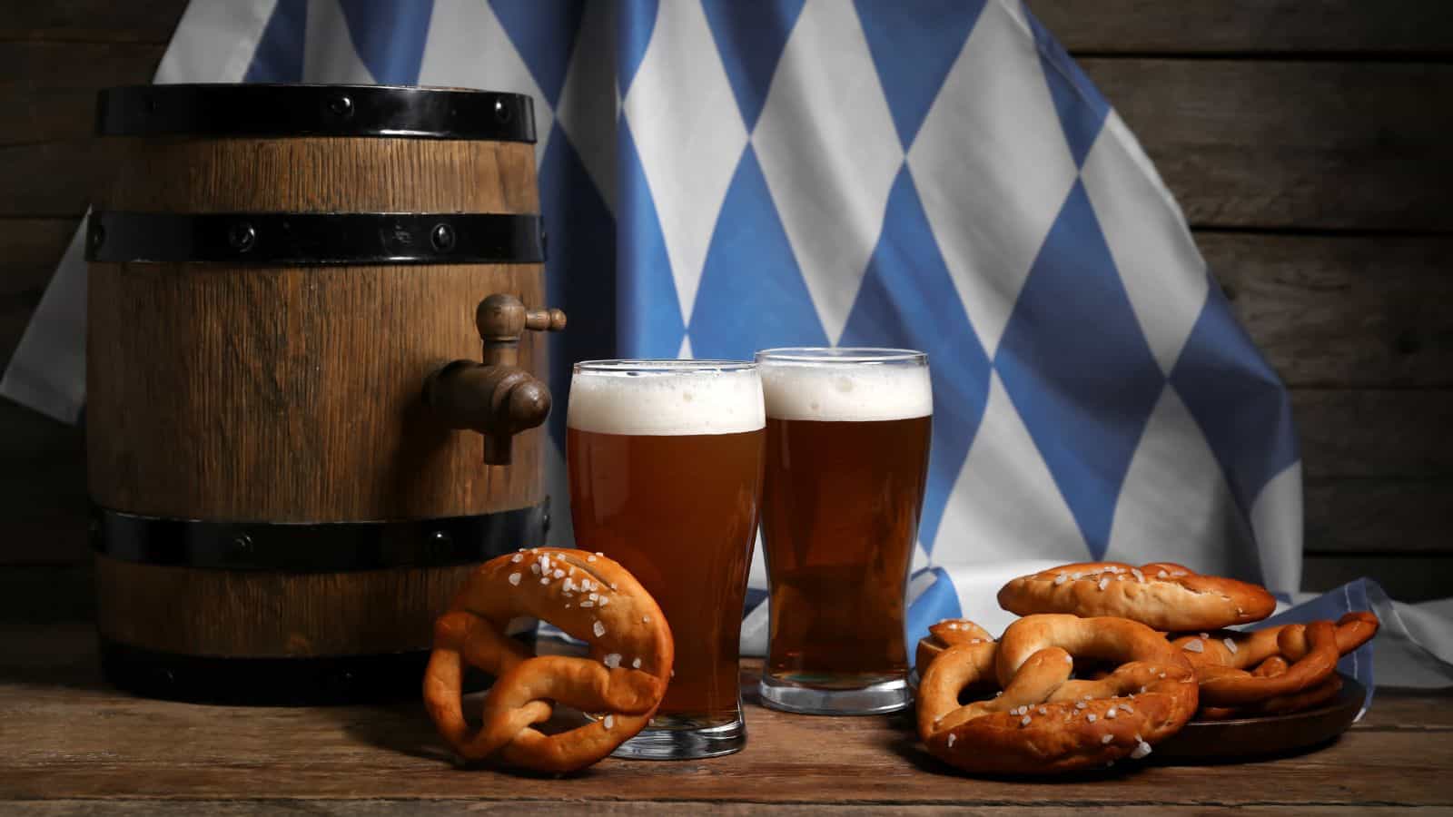 Two glasses of beer sit on a wooden table beside several pretzels. A wooden beer barrel is on the left. A blue and white checkered flag in the background suggests a Bavarian theme.