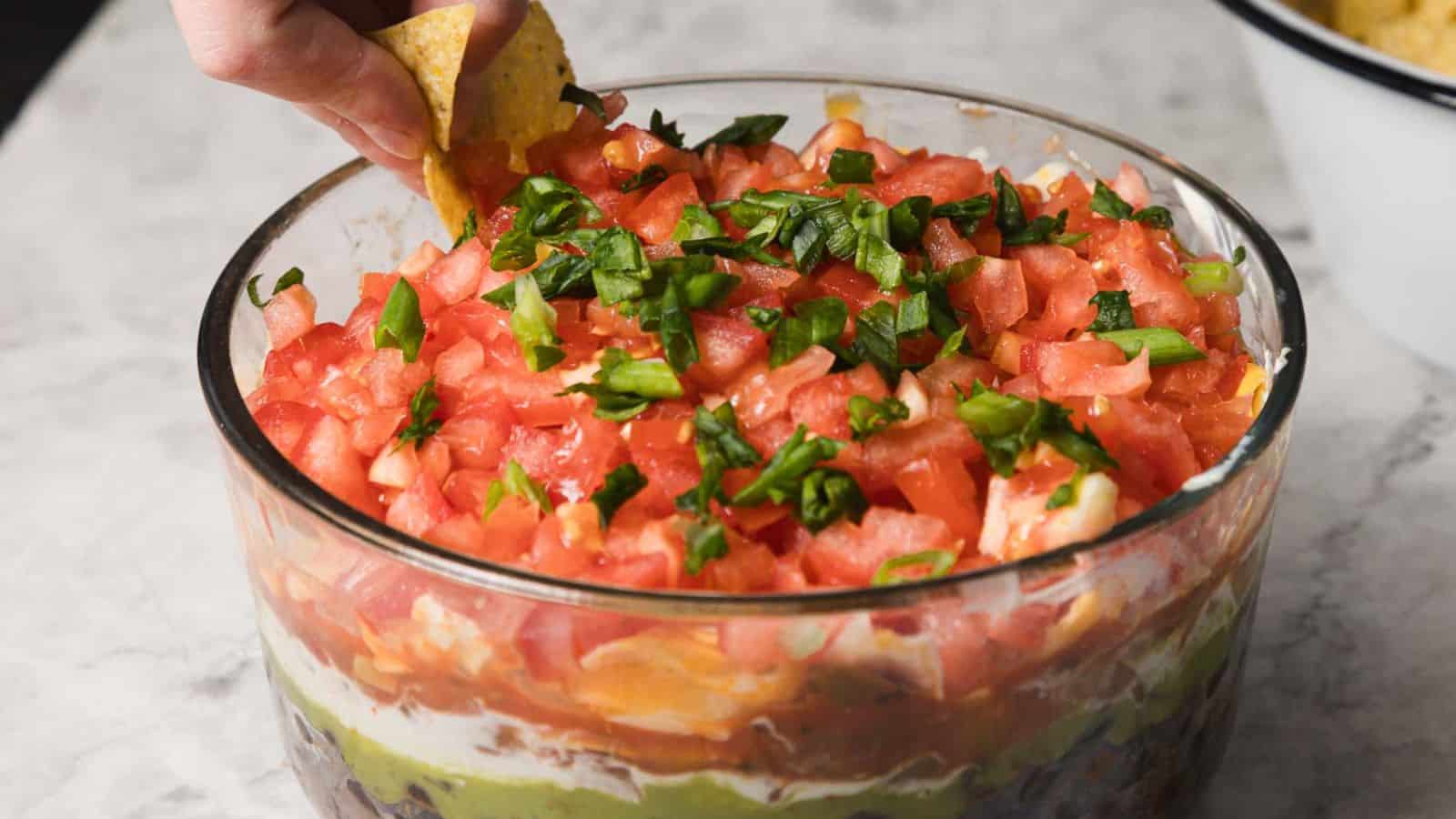 A hand holding a tortilla chip is dipping into a layered taco dip in a glass bowl. The dip includes layers of refried beans, guacamole, sour cream, shredded cheese, salsa, diced tomatoes, and chopped green onions on top.