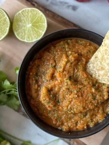 A bowl of salsa is placed on a wooden board. The salsa is garnished with a tortilla chip. Nearby are two lime halves and a bunch of cilantro. A purple cloth is partially visible in the background.