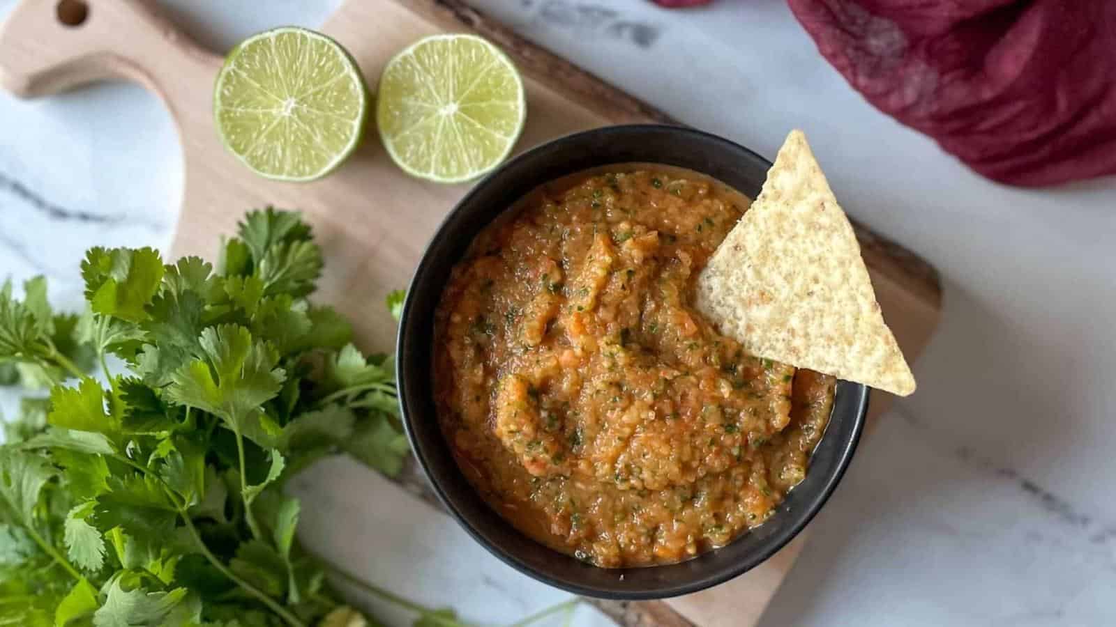 A bowl of salsa is placed on a wooden board. The salsa is garnished with a tortilla chip. Nearby are two lime halves and a bunch of cilantro. A purple cloth is partially visible in the background.
