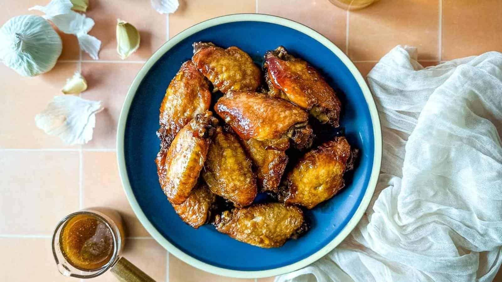 A blue bowl filled with glazed chicken wings placed on a tiled surface. Garlic cloves and a light-colored cloth are on the side, with a small jar of brown sauce in the corner.