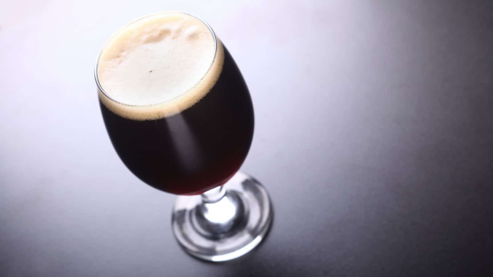 A glass of dark beer on a gray surface. The beer has a frothy, creamy head and is served in a stemmed glass, casting a shadow to the right.