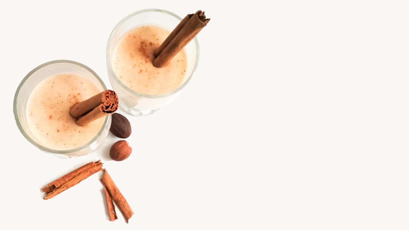 Two glasses of a creamy beverage topped with cinnamon sticks are placed on a light surface. Nearby, additional cinnamon sticks and whole nutmegs are scattered. The composition suggests a focus on flavor and ingredients.