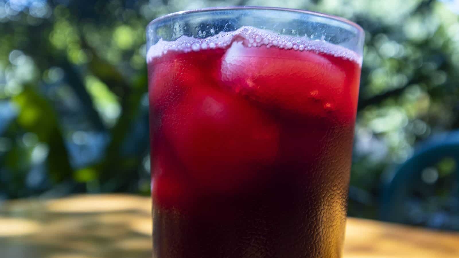 A glass filled with a deep red beverage and ice cubes. The background shows blurred greenery, suggesting an outdoor setting. The glass is placed on a wooden surface.