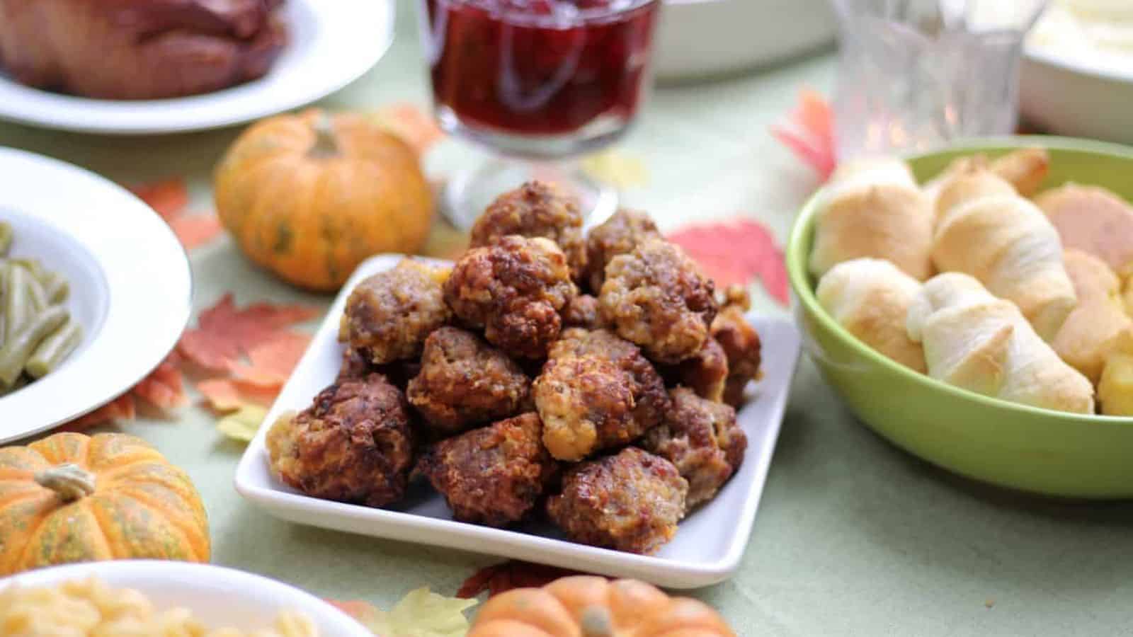 A table with a plate of meatballs in the center. Surrounding it are dishes with bread rolls, green beans, and a beverage in a glass. Small pumpkins are used as decorations on the table, which has a festive fall theme.