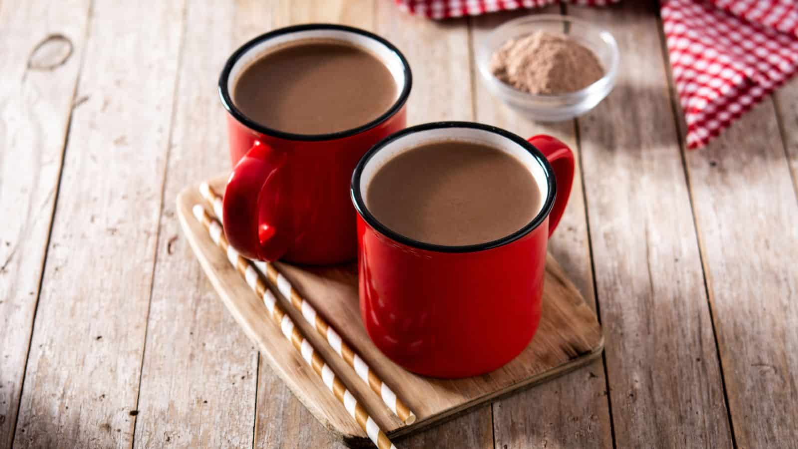 Two red mugs filled with hot chocolate are placed on a wooden tray. The tray rests on a rustic wooden table. Beside the mugs are two striped straws, and in the background, there is a small bowl filled with cocoa powder and a red checkered cloth.