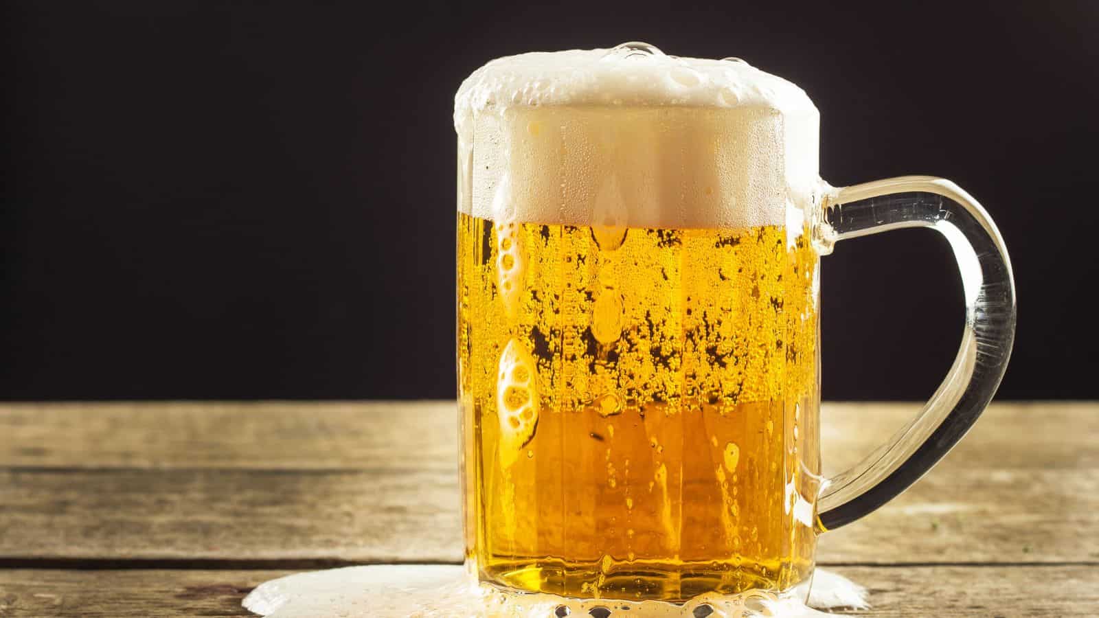 A glass mug filled with frothy beer sits on a wooden surface. The beer is golden and bubbly, with foam spilling over the rim. The background is dark, highlighting the vividly illuminated beverage.