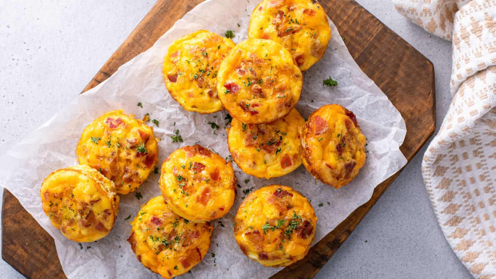 A wooden board holds a piece of parchment paper with ten baked egg muffins. The muffins are golden brown with visible bits of bacon and herbs sprinkled on top. A beige and white checkered cloth is partially visible on the side.