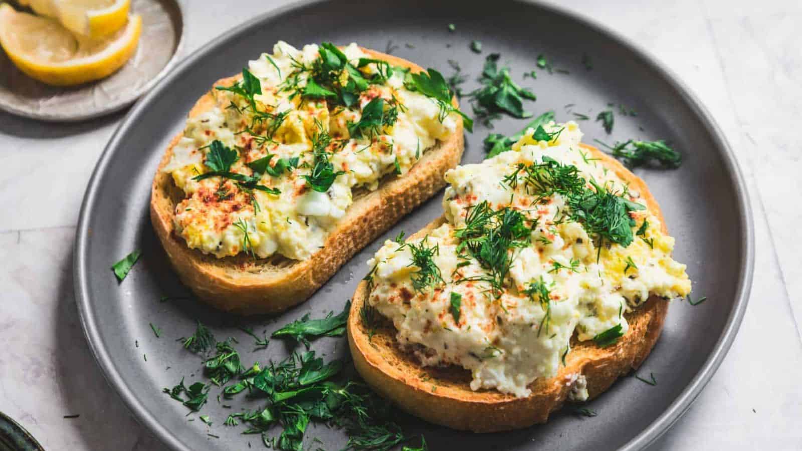 Two slices of bread topped with a creamy egg salad, garnished with chopped herbs, rest on a gray plate. A few lemon wedges are placed on a small dish in the background. The scene is set on a light-colored surface.
