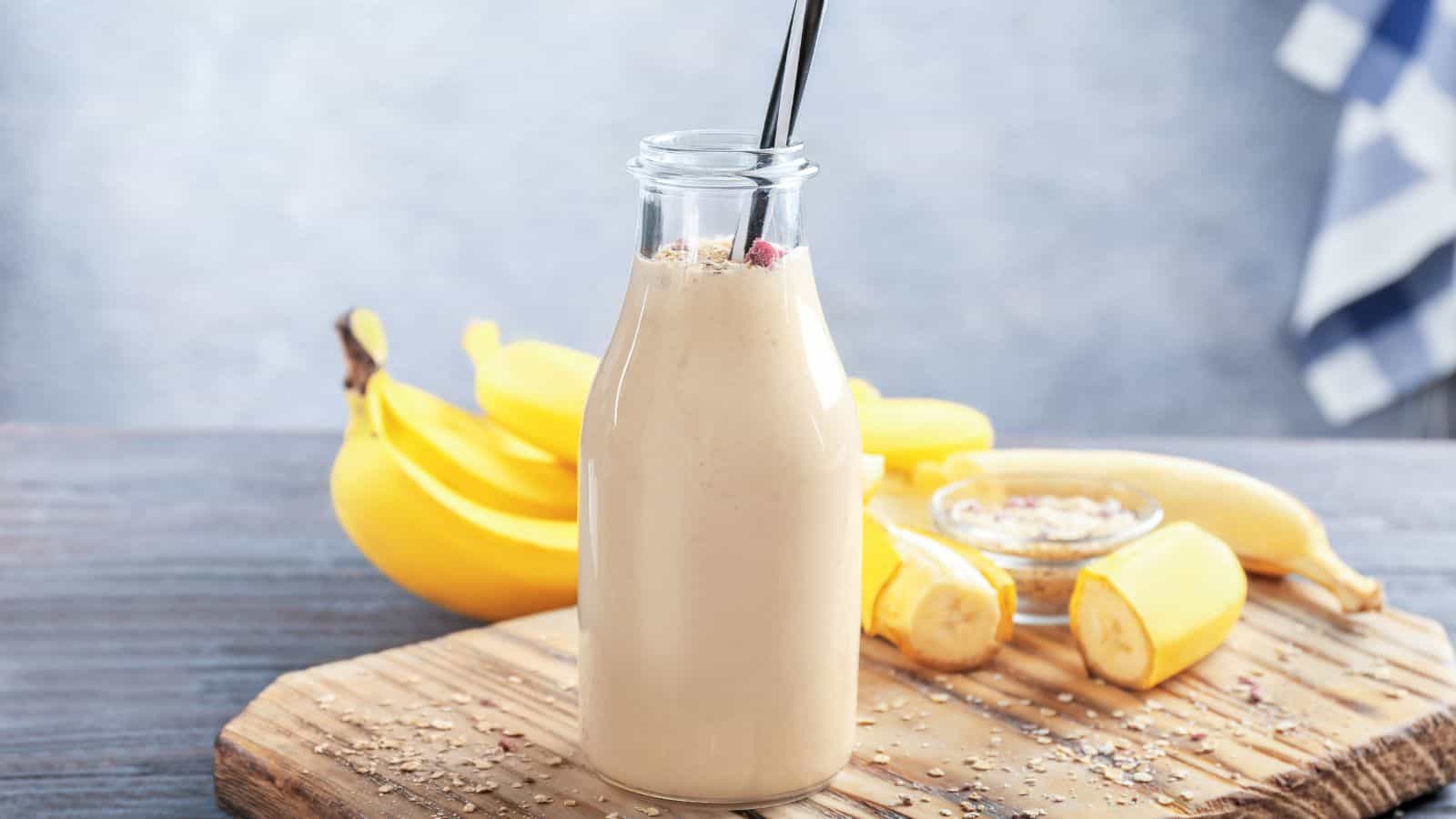 A glass bottle filled with a creamy smoothie, garnished with oats, on a wooden board. Two black straws extend from the bottle. In the background, there are whole bananas, a sliced banana, and a small bowl of oats.