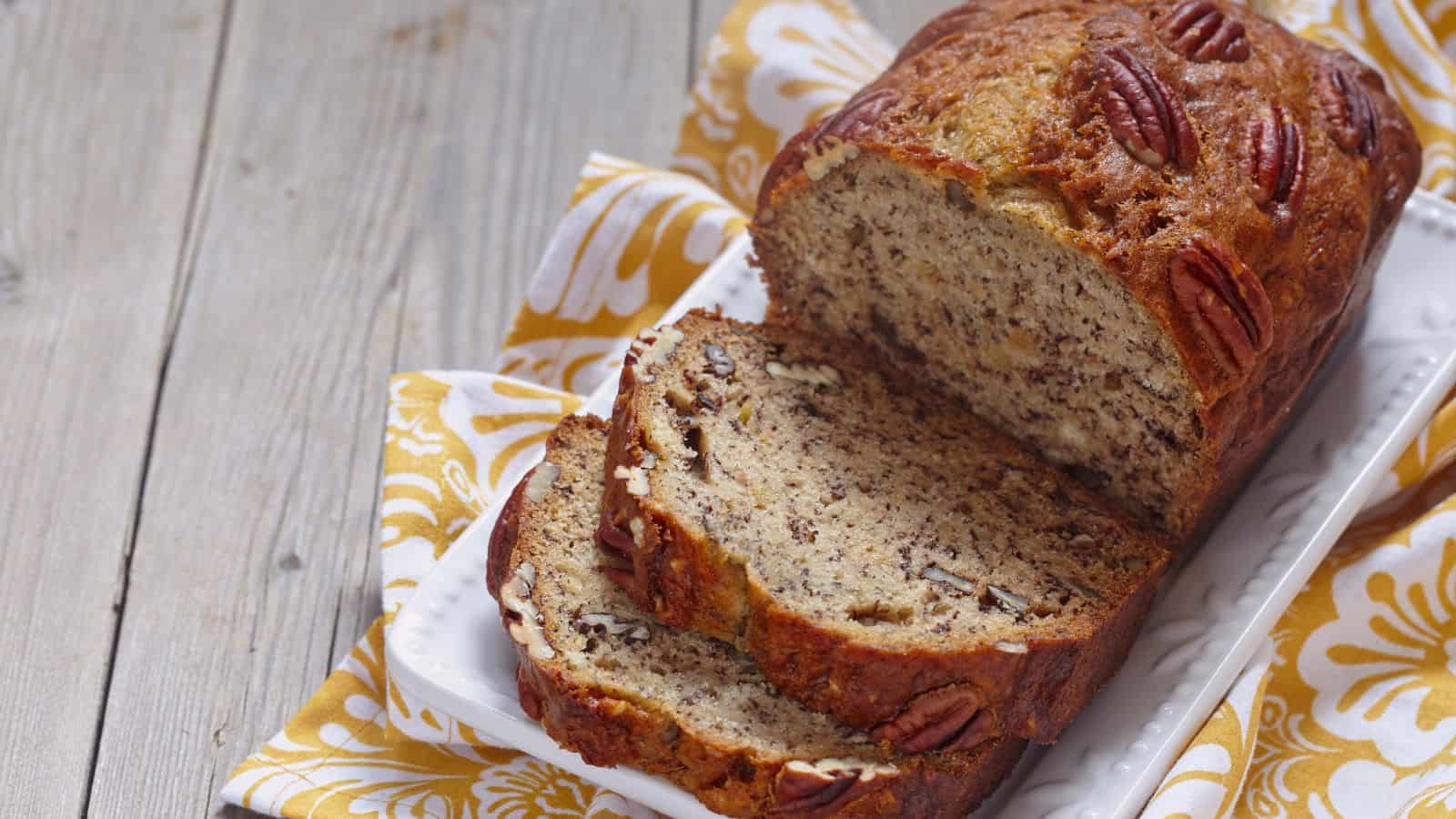 A loaf of banana bread with pecans sits on a rectangular white plate. Two slices are cut from the loaf. The plate rests on a yellow and white patterned cloth. The background surface is light gray wood.