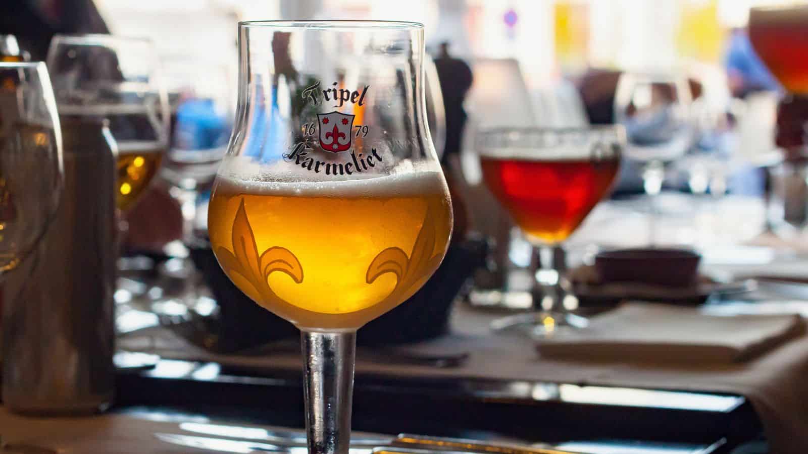 A glass of Tripel Karmeliet beer sits on a table set for dining. The glass is filled with a light amber beer and has a decorative design. Other glasses and tableware are visible in the background, with a soft focus effect.