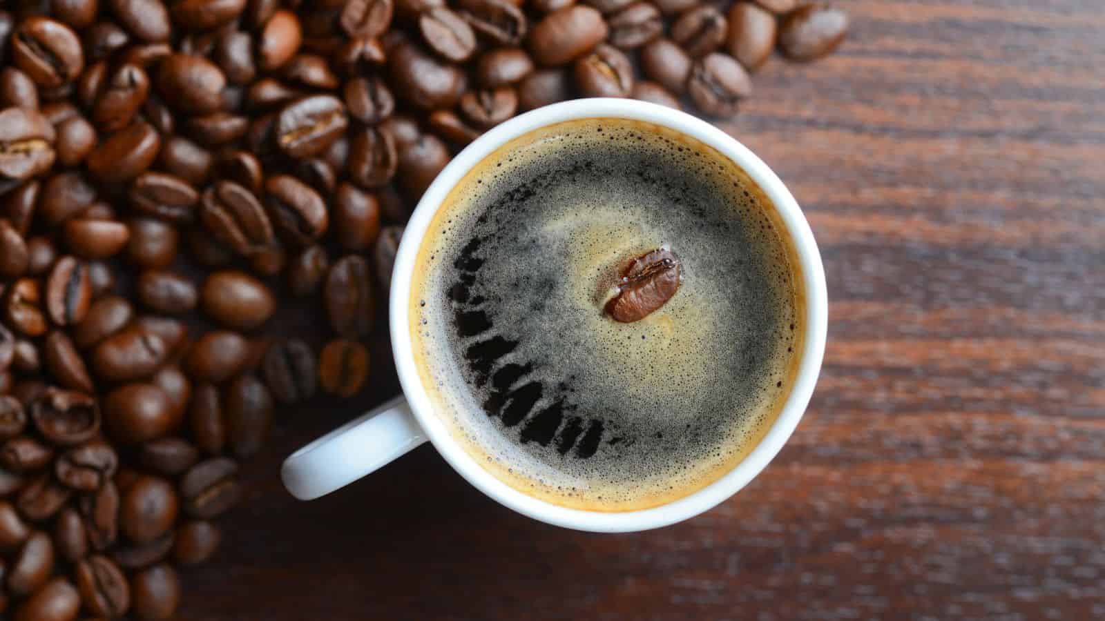 A white ceramic mug filled with black coffee on a wooden table. A single coffee bean floats on the surface. A pile of roasted coffee beans is scattered to the left of the mug.