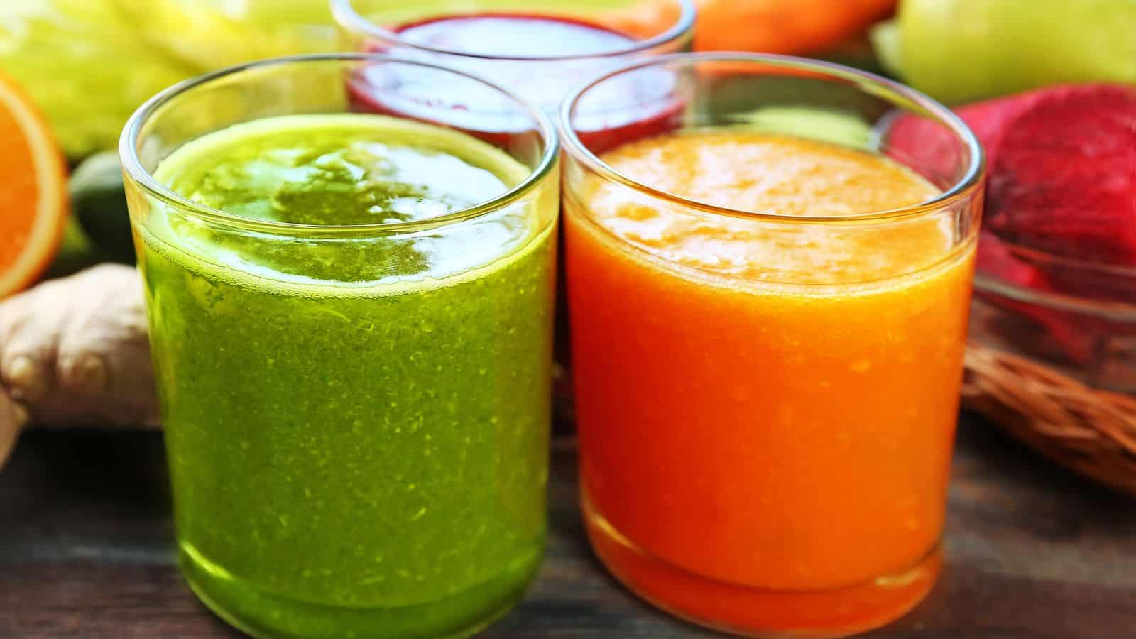 Two glasses of freshly blended smoothies sit side by side. One glass contains a bright green smoothie, and the other has an orange smoothie. The background features fresh fruits and vegetables, slightly out of focus.