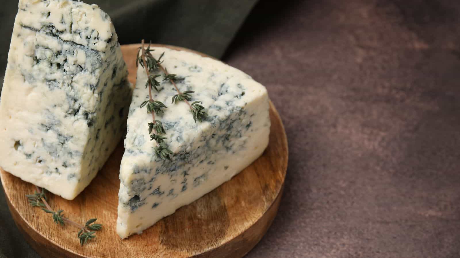 Two wedges of blue cheese are placed on a round wooden board. The cheese has visible blue veins. A sprig of thyme rests on one wedge. The background is a dark textured surface.