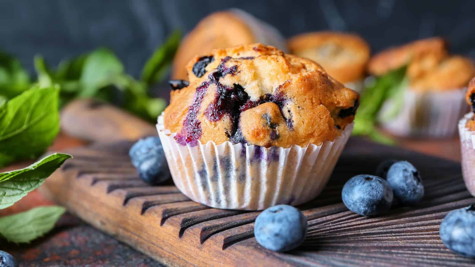 A blueberry muffin in a paper liner is placed on a wooden board. Fresh blueberries and green leaves are scattered around. More muffins and an indistinct background complete the scene.