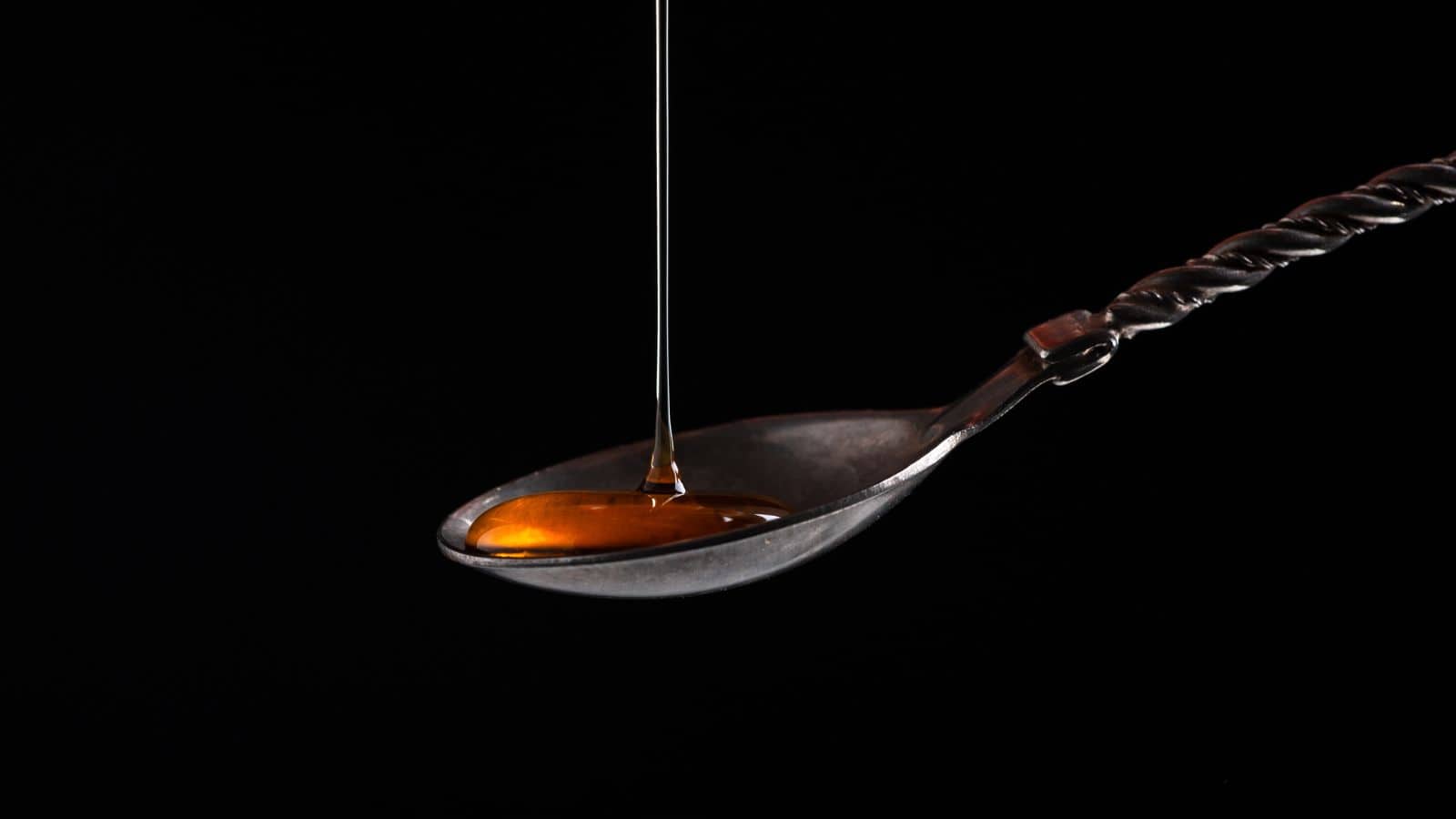A stream of golden liquid pours into an old-fashioned metal spoon with a twisted handle, set against a black background.