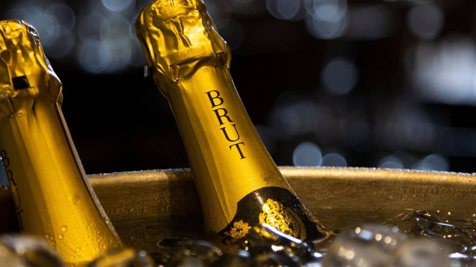 Close-up of a champagne bottle labeled "Brut" with gold foil wrapping. The bottle is immersed in ice inside a bucket, with blurred sparkling lights in the dark background.