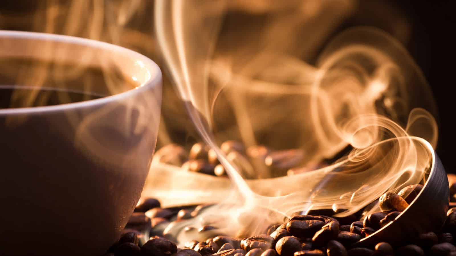 A steaming cup of coffee sits among scattered coffee beans. Wisps of steam rise dramatically, creating swirling patterns in the warm light. The close-up highlights the rich texture of the beans and the inviting aroma of the coffee.