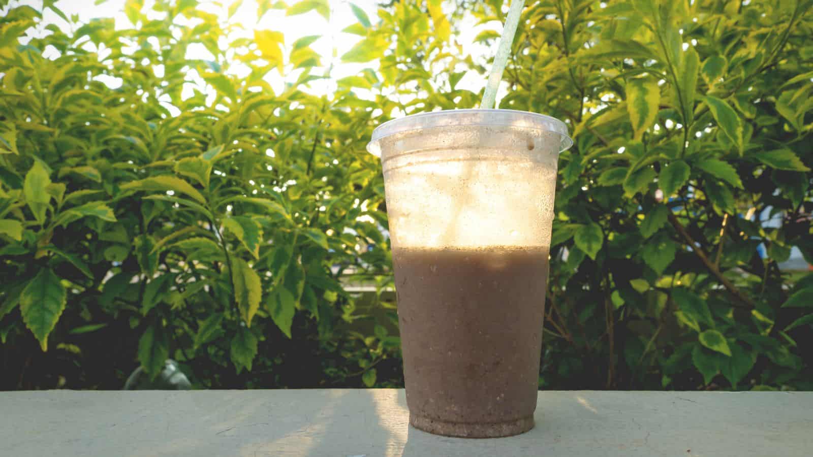 A plastic cup with a dome lid and straw contains a brown smoothie. It is placed on a flat surface outdoors, with green foliage in the background, illuminated by sunlight.